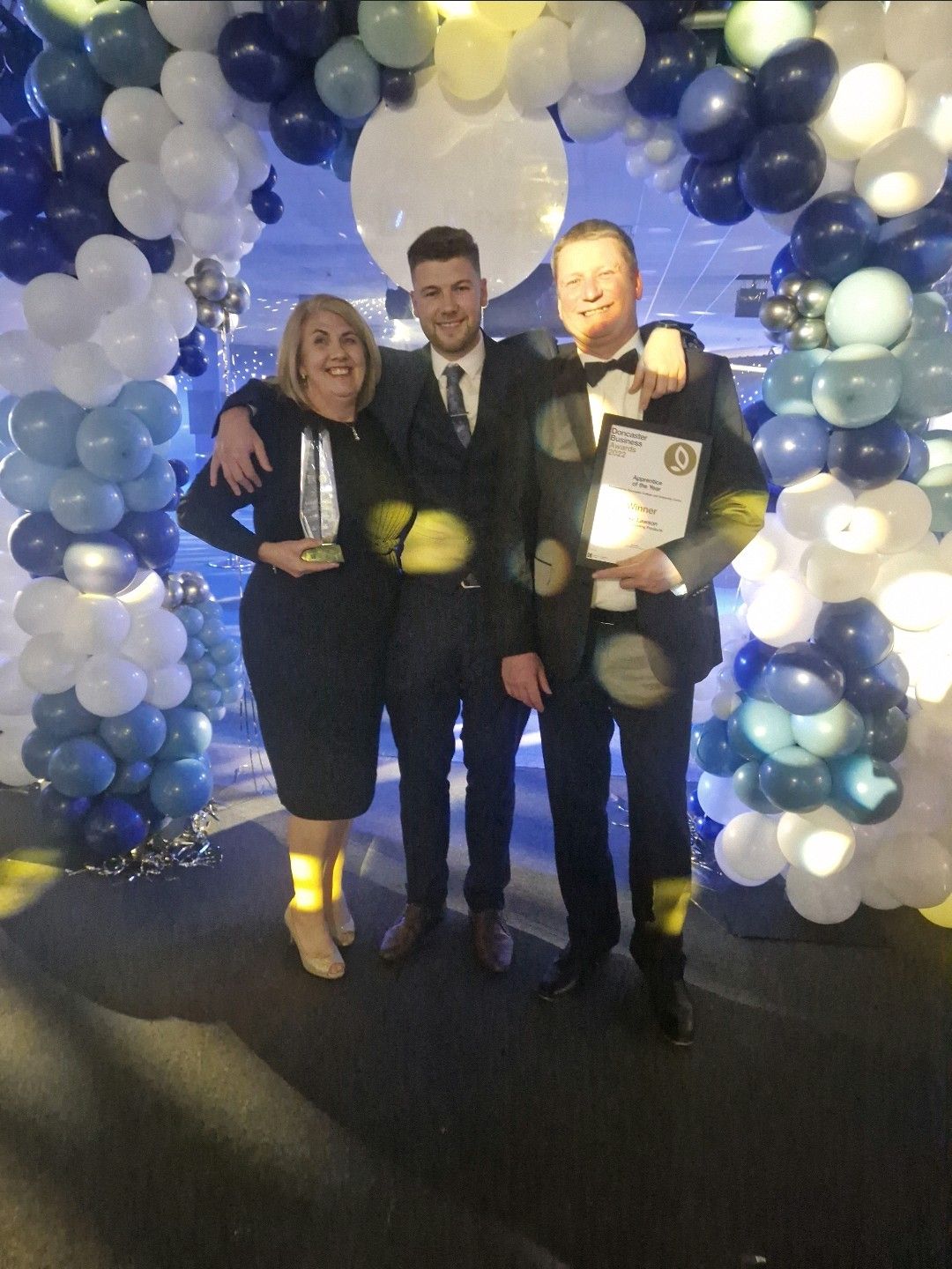 Automation Engineer Apprentice Luke Lawson is pictured holding his Apprentice of the Year Award at the 2022 Doncaster Business Awards. Stood to his left is his Mum Joanne, and to his left his Dad Alan. They are beneath a blue and white balloon arch.